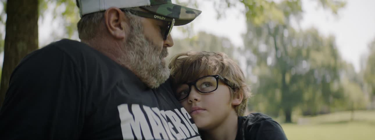 A boy rests his head against his dad as they sit outside near the trees