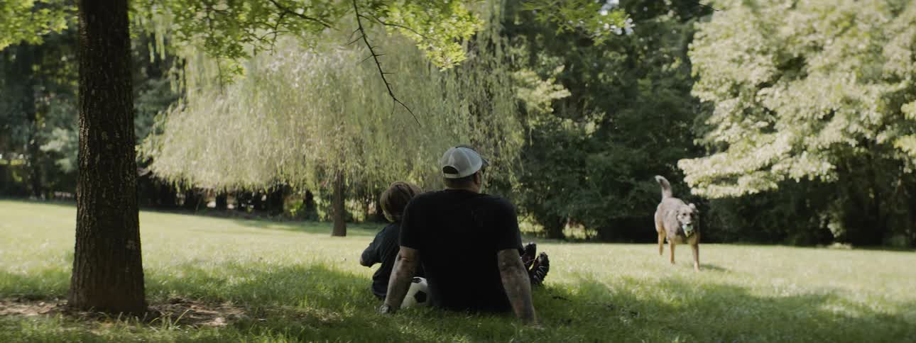 Two people sit in the grass as a dog retrieves a ball