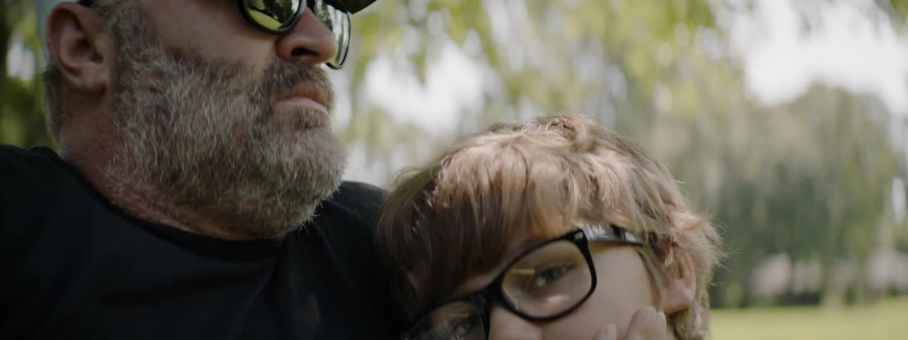 A boy wearing glasses lays his head on his father while in a park