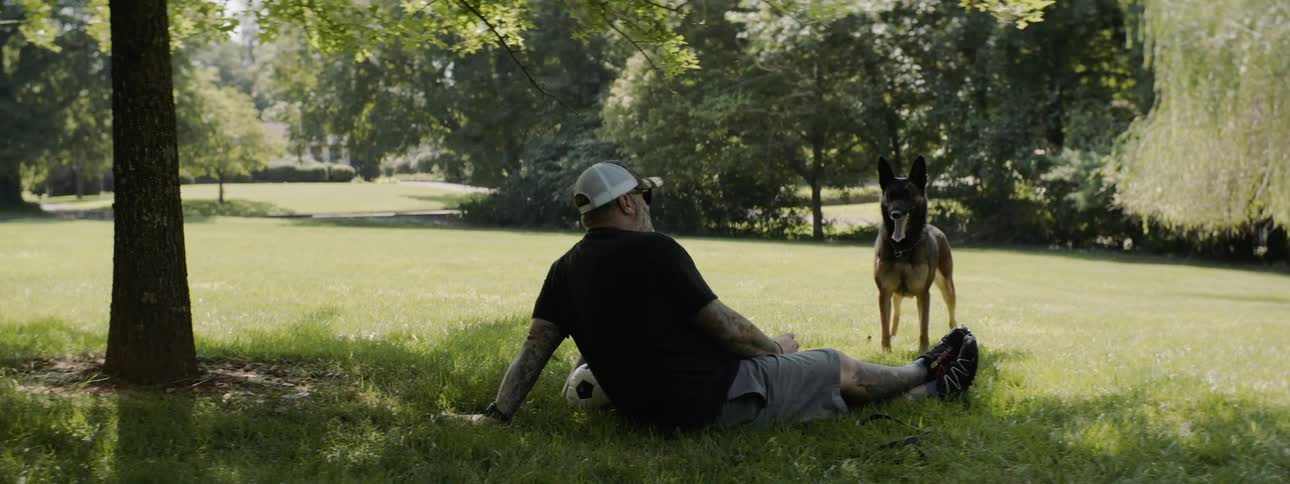 Dog stands wagging tail next to two people sitting on grass