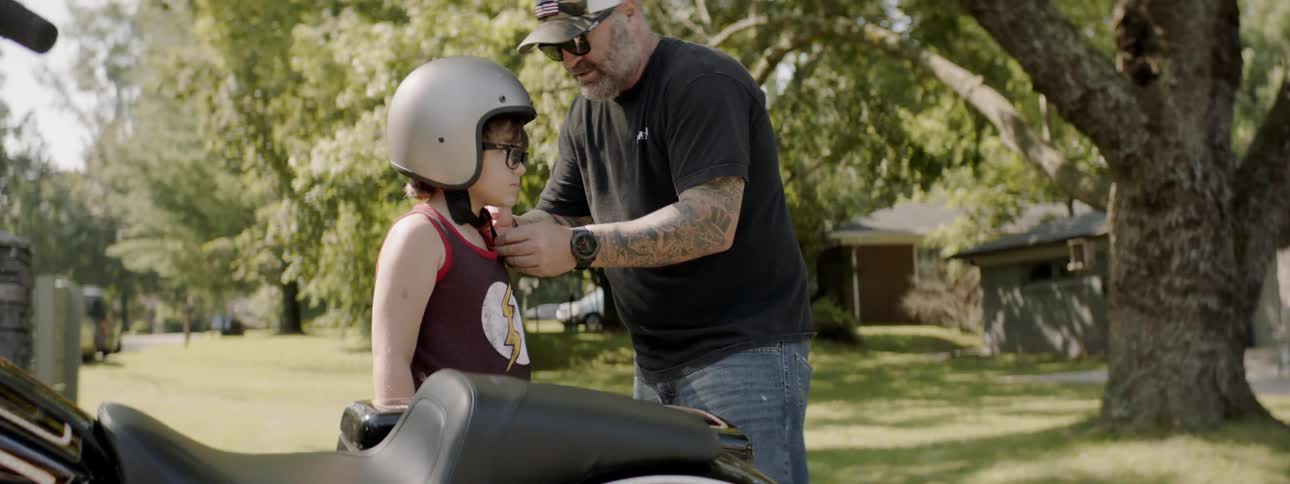 Man wearing cap and sunglasses makes boy wear helmet in the countryside on a sunny day