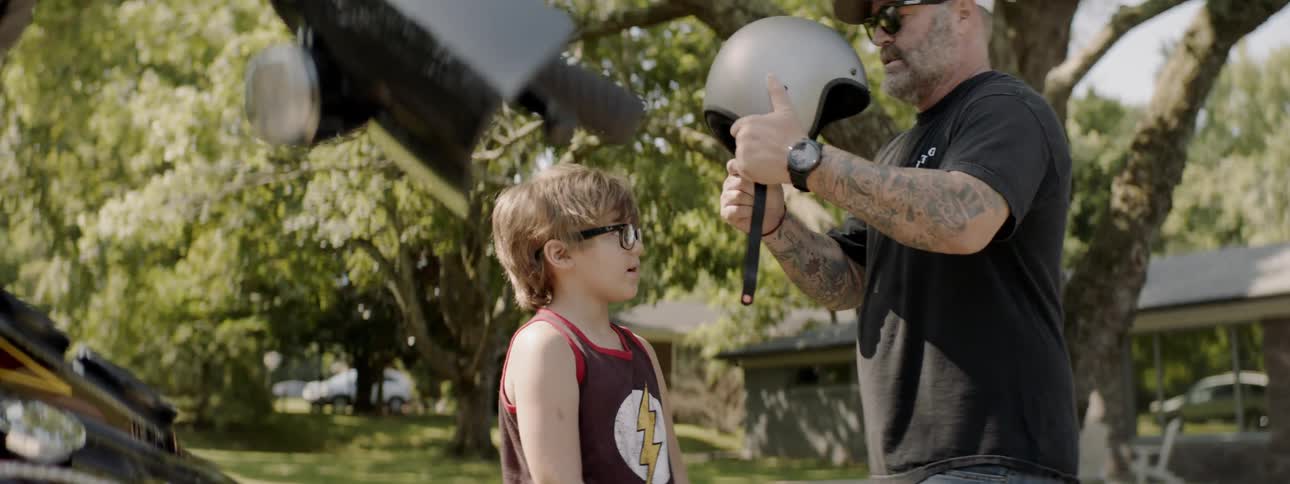 Man wearing white cap and sunglasses makes child wear motorcycle helmet in the countryside during daylight