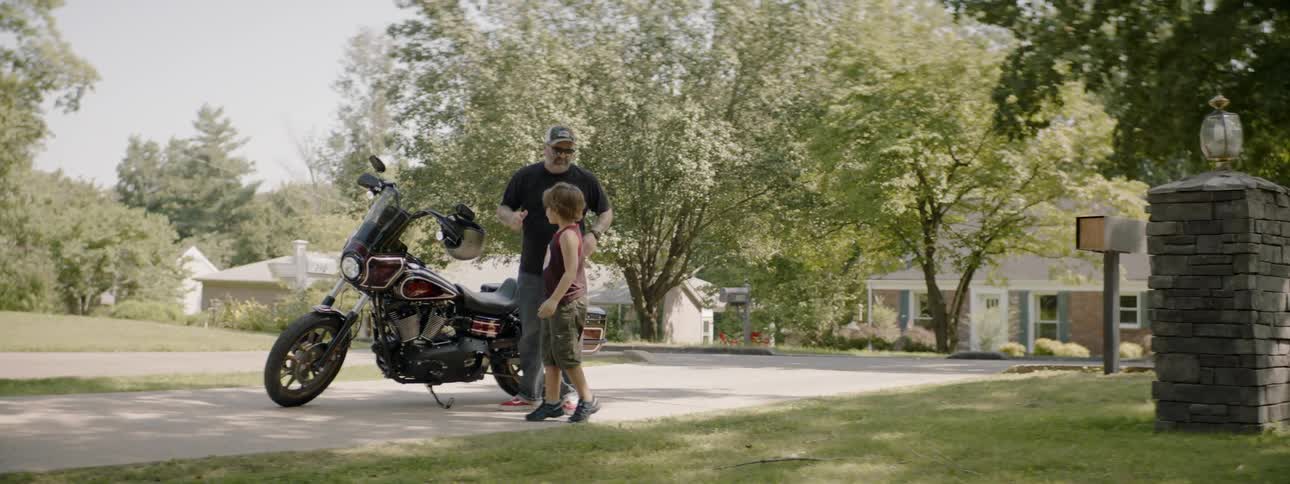 A man takes a motorcycle helmet off a boy and hangs the helmet on the handlebar of a motorcycle