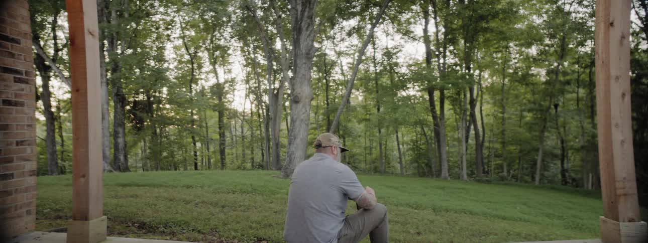 Bearded man sits in front of house on the grass with the view of forest during daylight