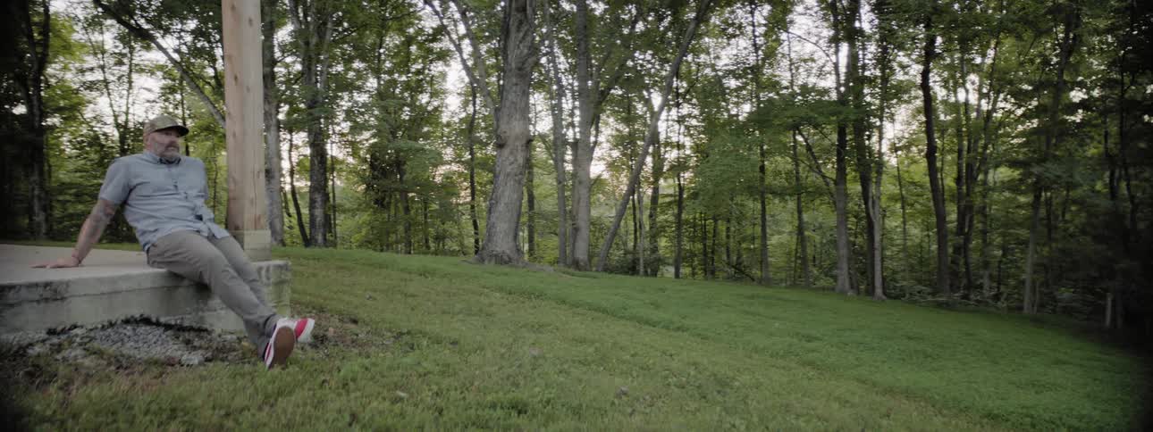 A burly and bearded man sits outside on a concrete slab in a grove of trees