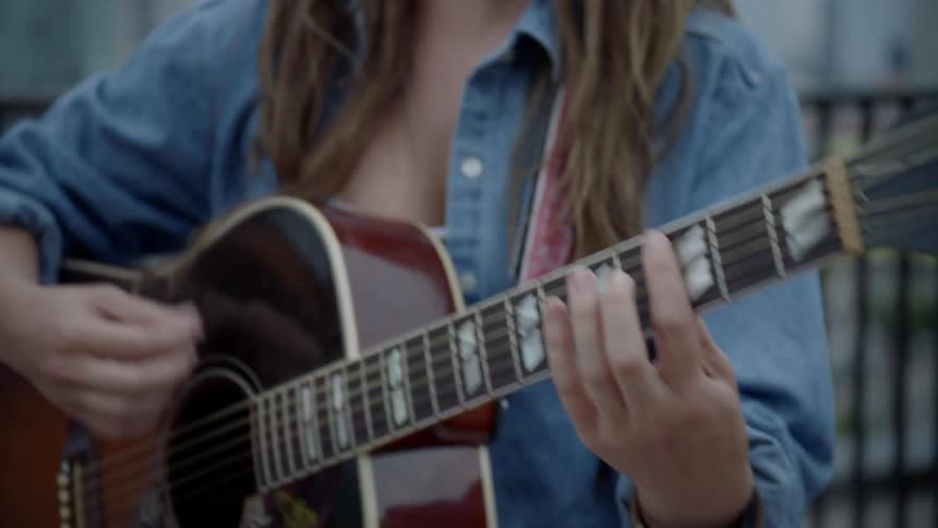 A young woman in a denim shirt is standing outside on the rooftop of a tall city building where she's happily singing and strumming her acoustic guitar