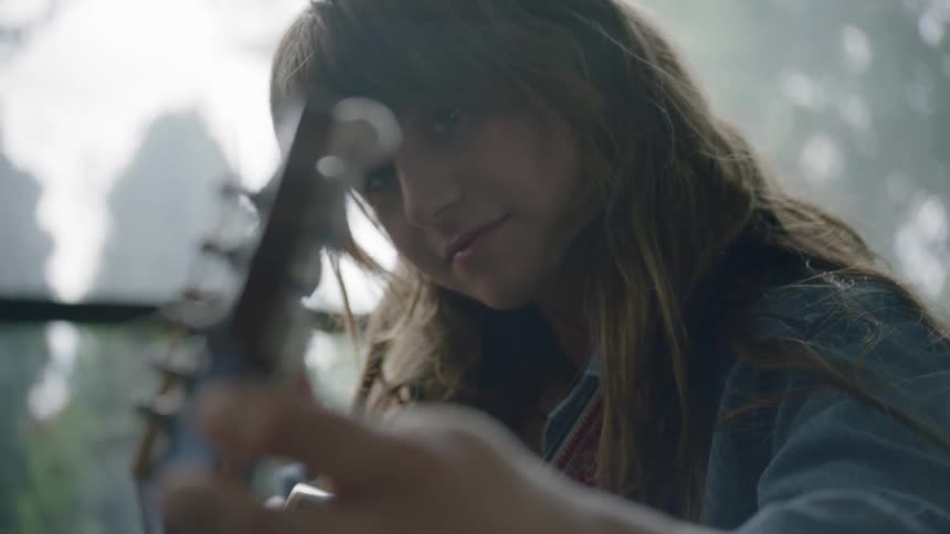 A girl smiles as she tunes her guitar while sitting in front of a window