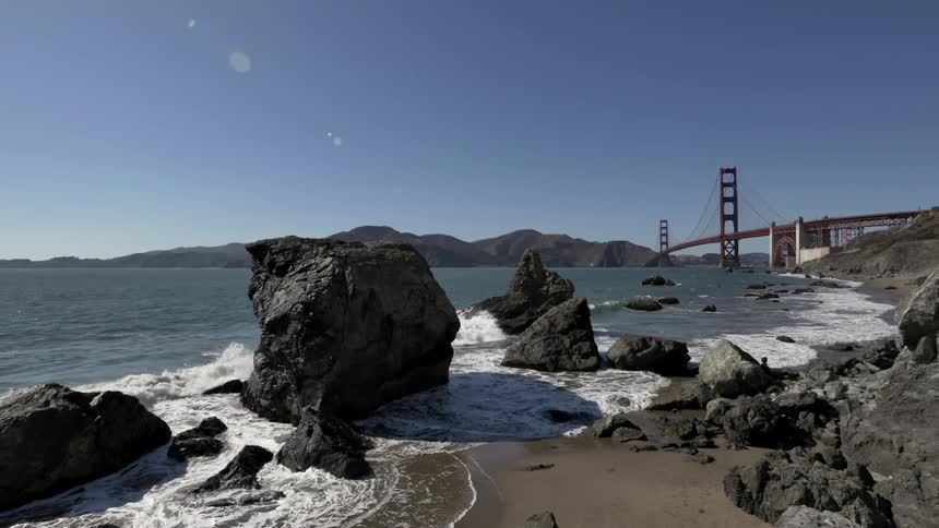 Waves foam and crash against rocks on the shoreline near a bridge