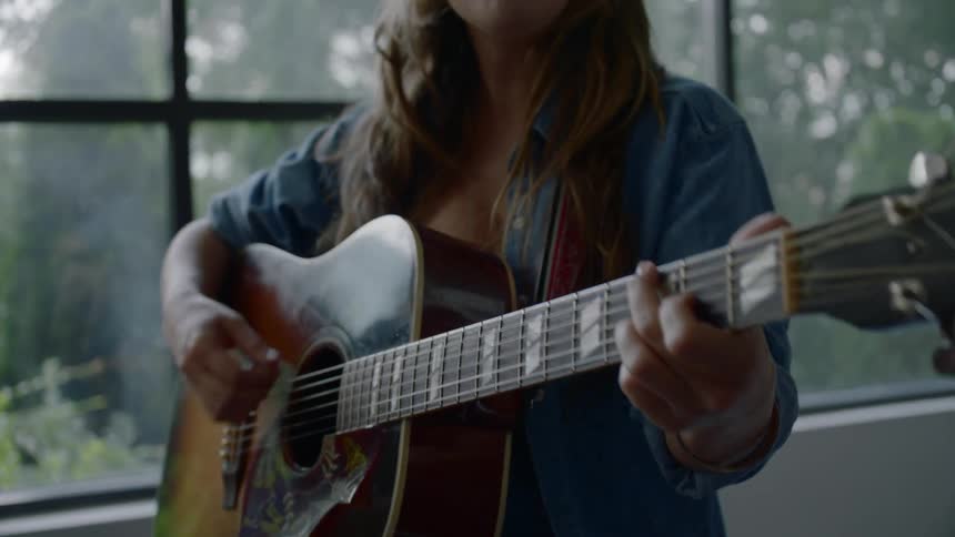 A white female stands near a wall of windows and sings while playing an acoustic guitar