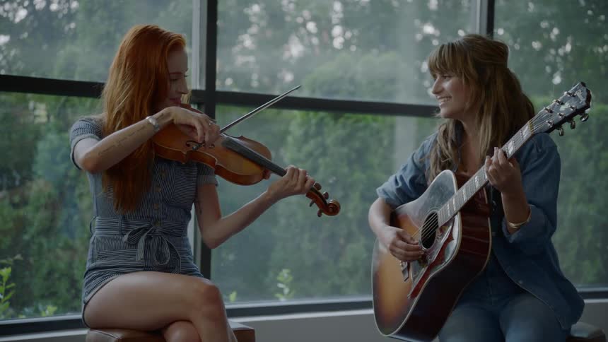 A woman with red hair plays a violin while a woman with brown hair plays a guitar