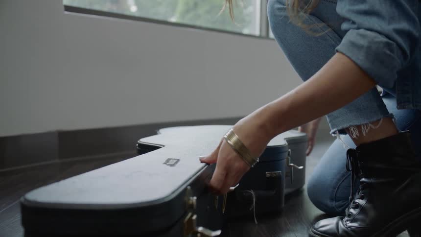 A white woman brings in a guitar case sets it down and takes the guitar out