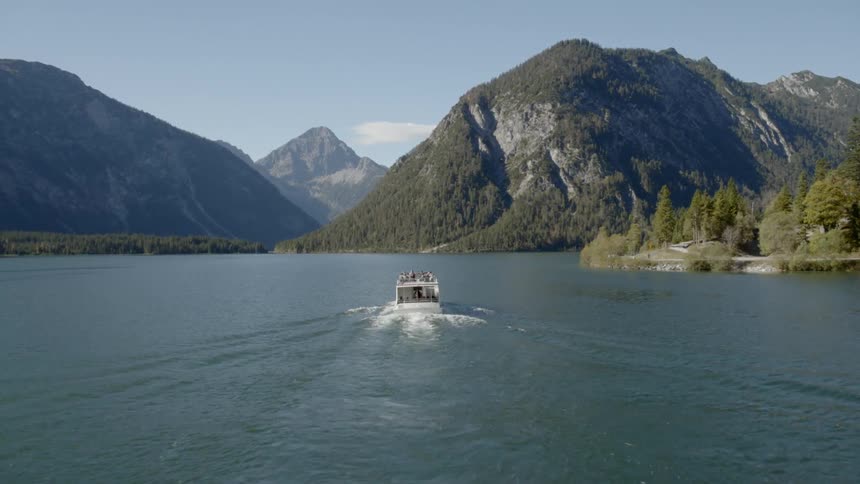 A touring boat motors along a large body of water that is surrounded by multiple large mountains and forests