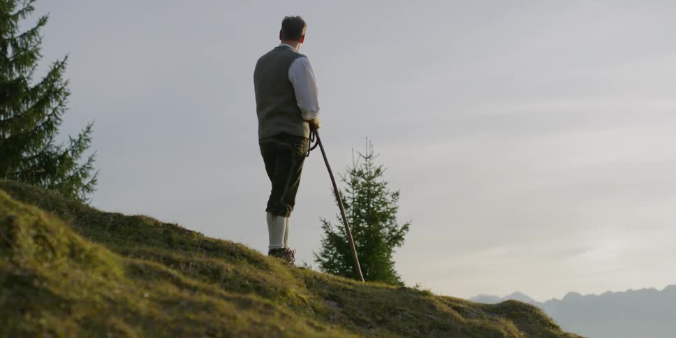 A man in hiking gear stands atop a grassy hill while holding a rope and a walking stick in a hilly and forested area