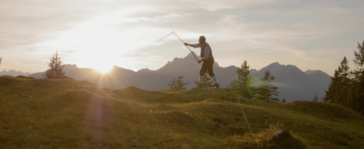 A man swings a whip on top of a hill with mountains behind him
