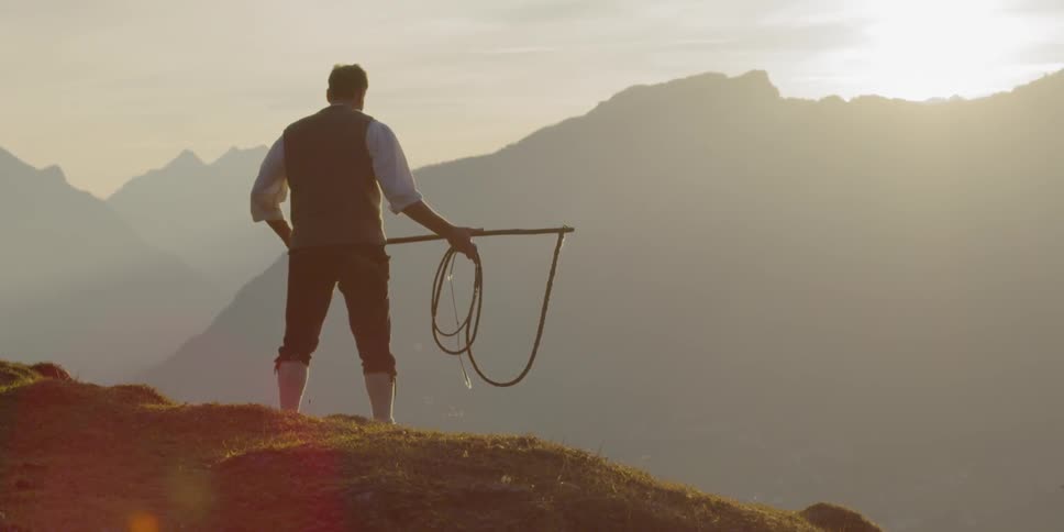 A stout man stands on a mountainside holding a large pole with a whip attached to the end