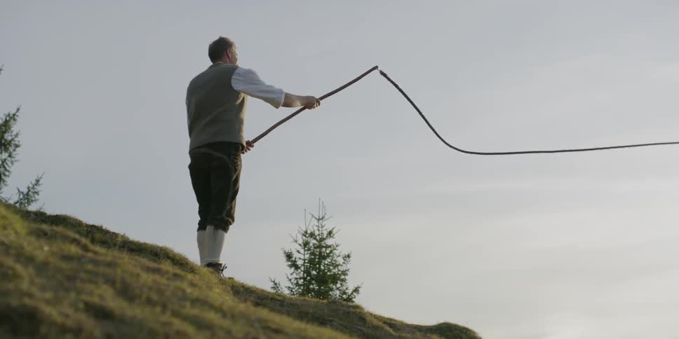 A white man stands on a hill swinging a whip attached to a long stick
