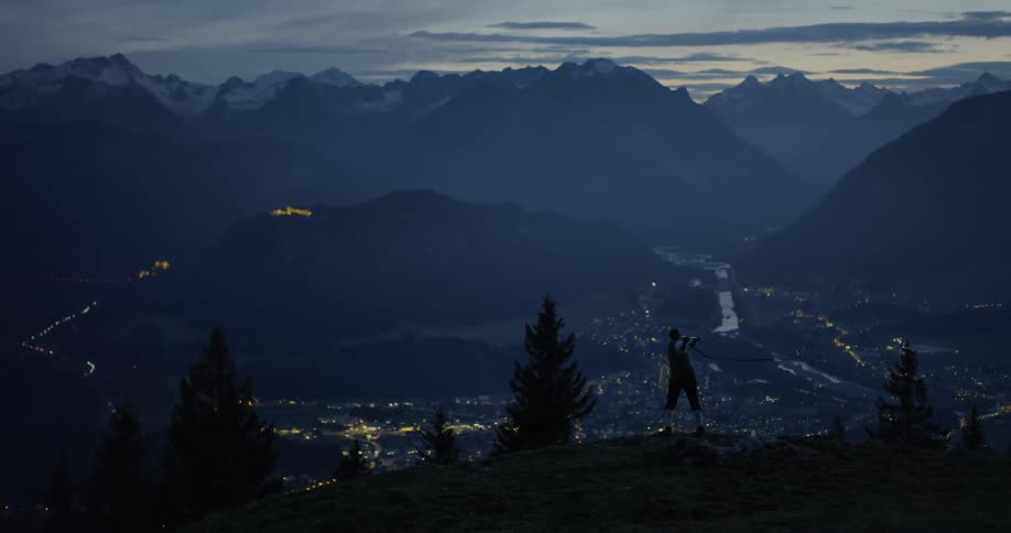 Man swings whip on high mountain with the view of illuminated town at dusk