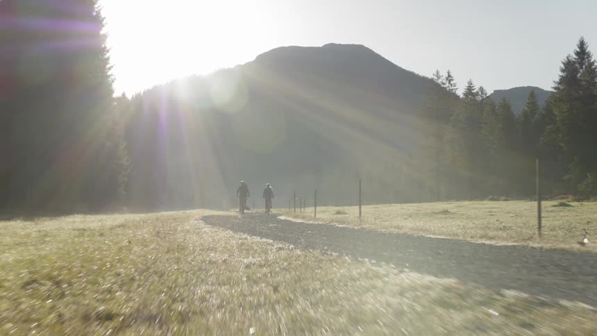 Two people ride bikes on a dirt road heading away from hills on a sunny day