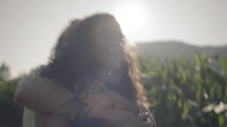 A young man playfully hugs and wrestles a young woman with curly hair as they walk along a path in a cornfield