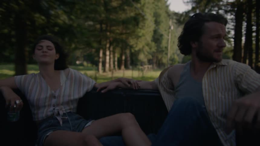 A man and a woman ride in the back of a pickup truck driving down a tree lined road