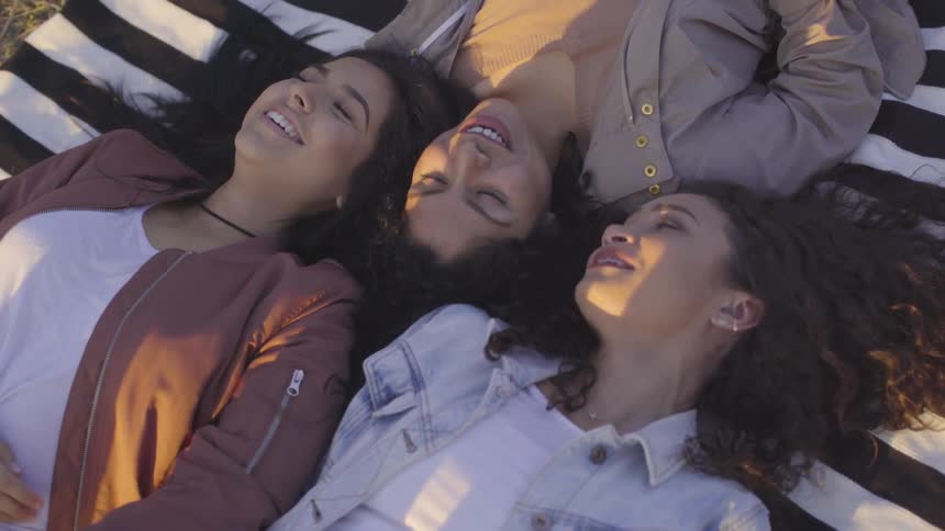 Three women chatting and laughing while laying together on a blanket outdoors