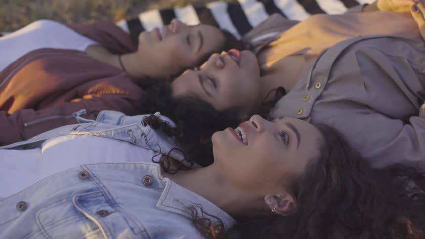 Three women lie on a blanket in the grass with their heads together looking up at the sky talking and smiling
