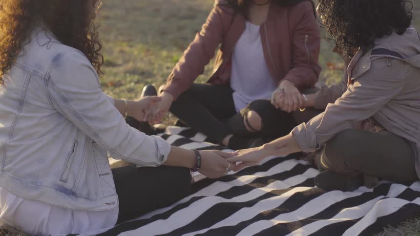 Three young woman sit on a blanket and hold hands