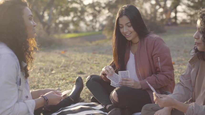 Three women play cards while sitting on a blanket in the grass