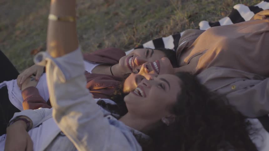 Three hispanic women lie on a black and white striped blanket laughing and taking a selfie