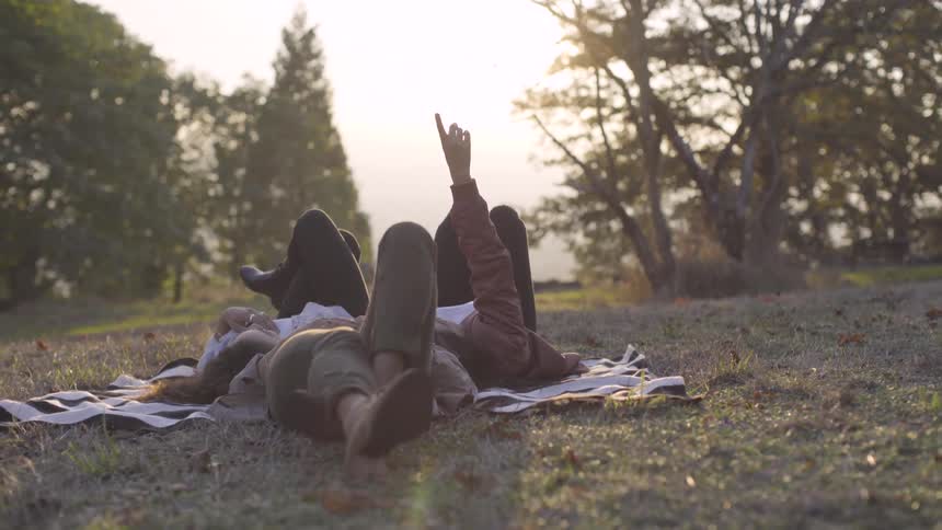 Friends look at the sky while laying on a blanket in a grassy field at sunset