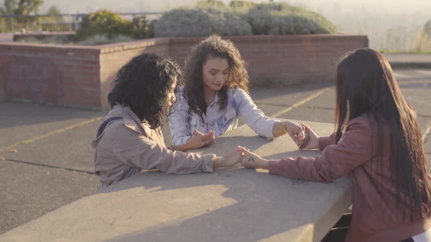 Three woman sitting outside on a table holding hands and praying