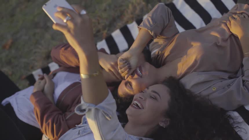 Three female friends smile and pose for a selfie while lying on a blanket on a lawn