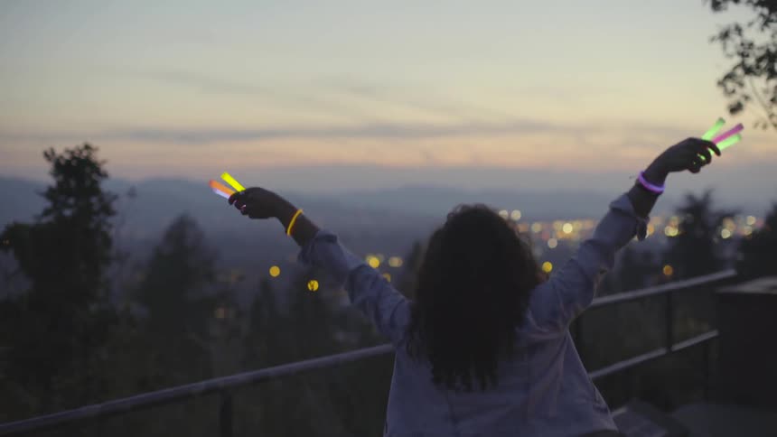 Woman stands outside at dusk waving glow sticks