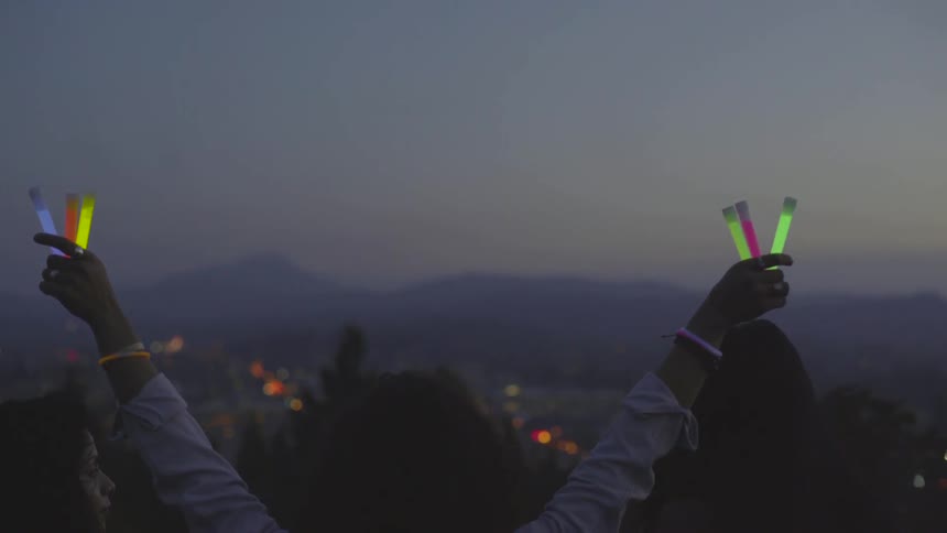 A woman lifts her arms into the air while holding glow sticks outside at dusk