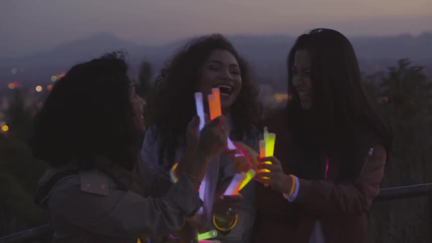 Three brunette women dance with light sticks on the hill across to the city at dusk