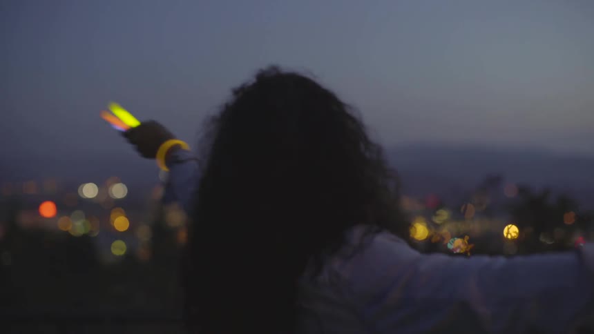 A woman smiles while spinning in circles with glow sticks outside at dusk