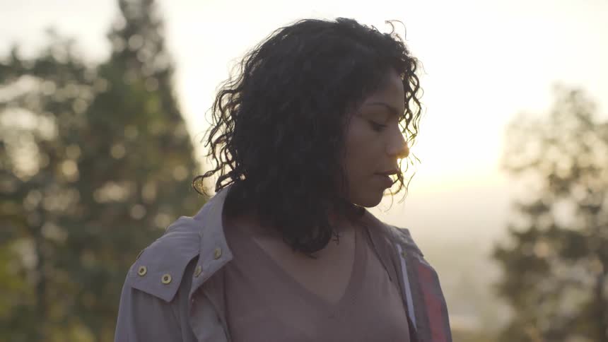 A woman with curly hair stands outside near trees at sunset
