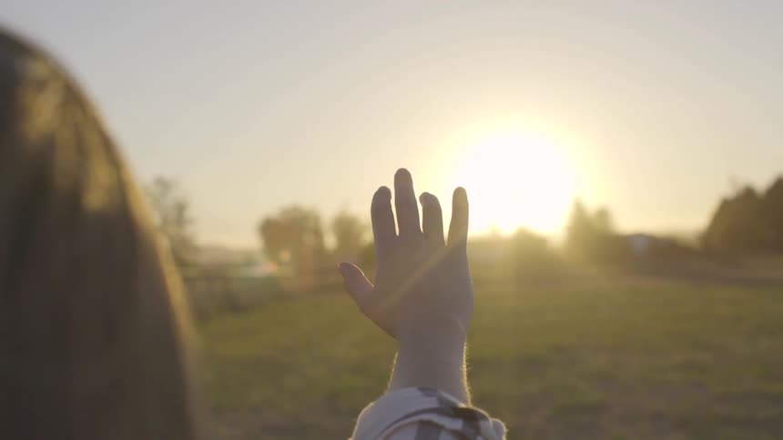 A woman stand on the grass and reaches her hand out to the sunlight