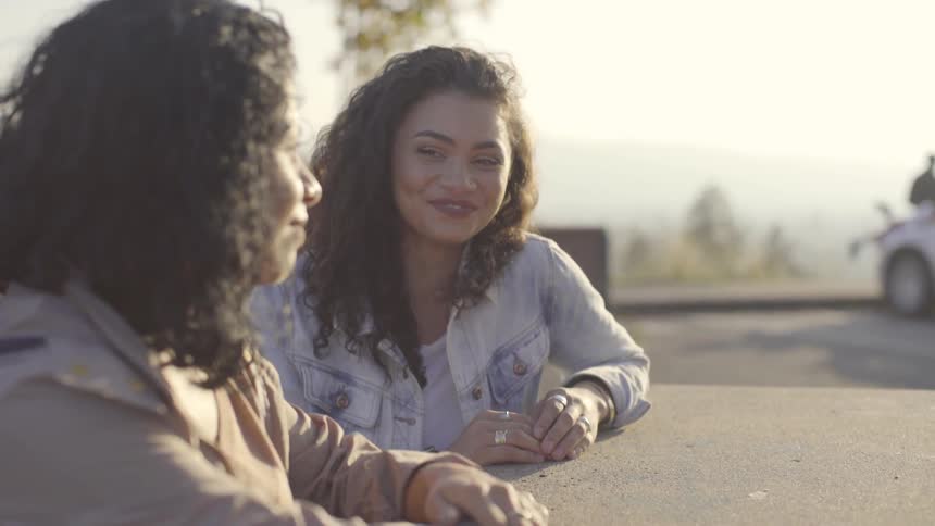 Two women sit at an outdoor table at an overlook and talk and smile on a sunny day