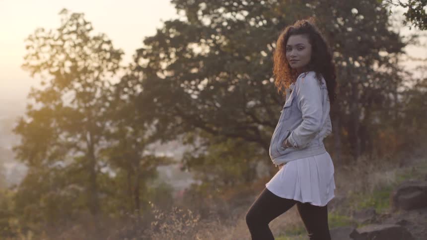 A woman stands with one foot on a stone and hands in pockets on a hill top at sunset