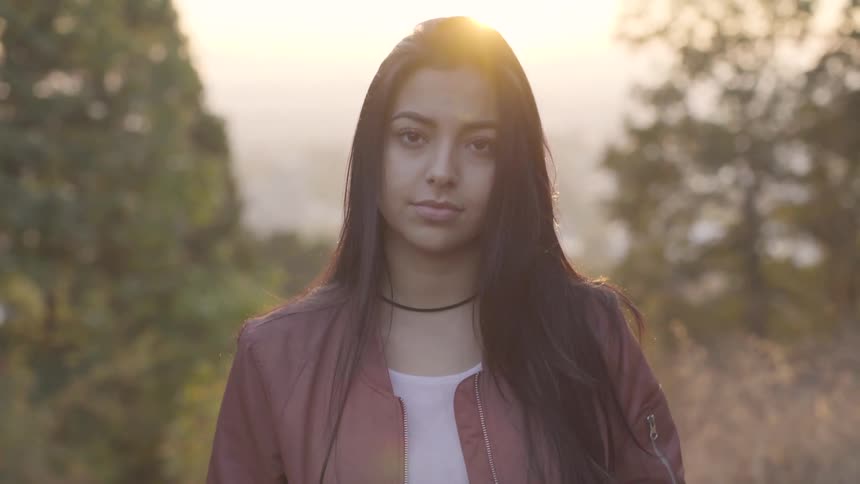 A woman with long hair bends her head then looks up with trees behind her at sunset