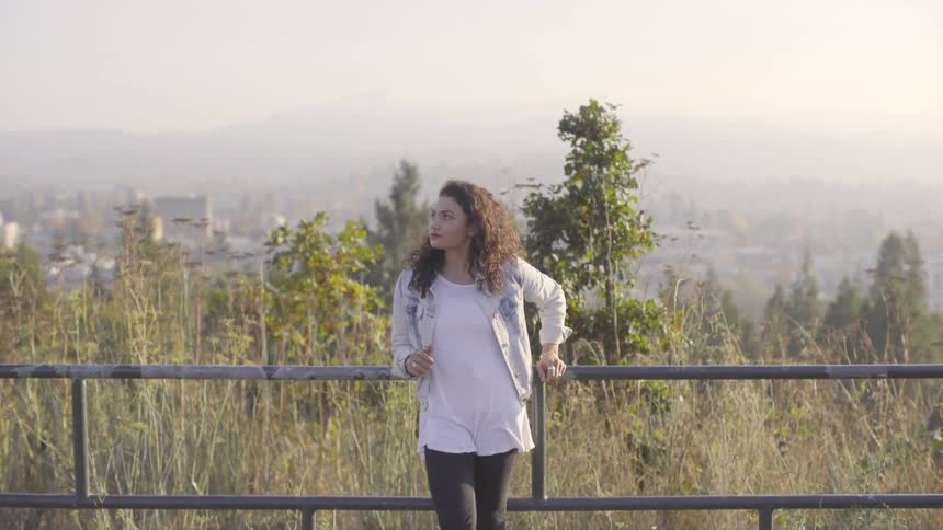 Woman stands against rail in front of grassy field and fog covered city