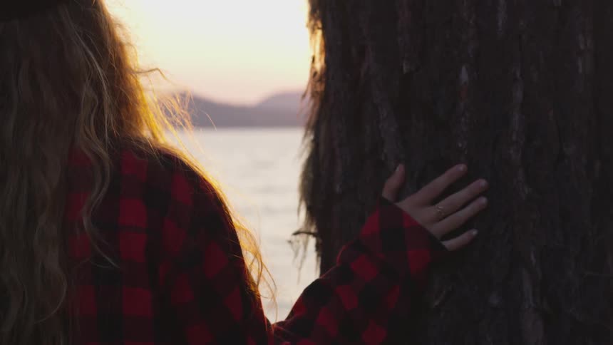 A woman wearing a flannel shirt touched the bark of a tree while looking at the water at sunrise