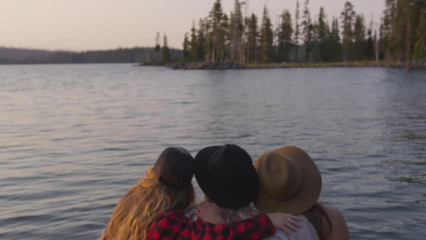 Three girl friends all wearing hats and with their arms around each other look out at the water