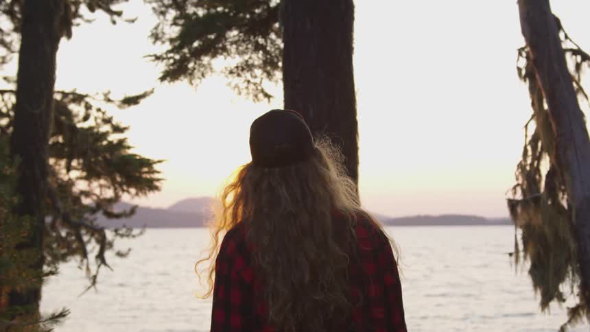Woman walks through trees toward the water at sunset