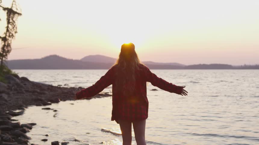 A woman raises her hands to the sky on the shore of a lake