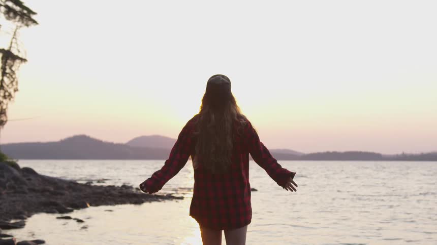 A woman stands on the shore and stretches out her arms then raises them over her head at sunset