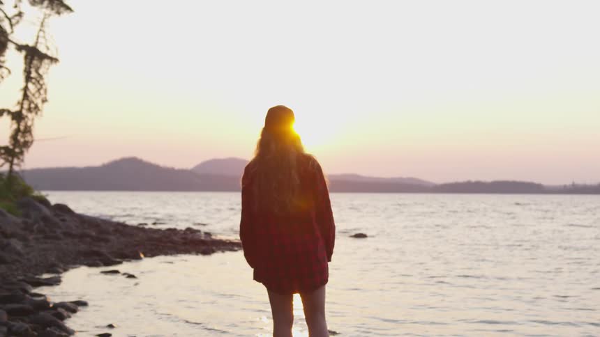 A woman stands on a rocky shore at sunset