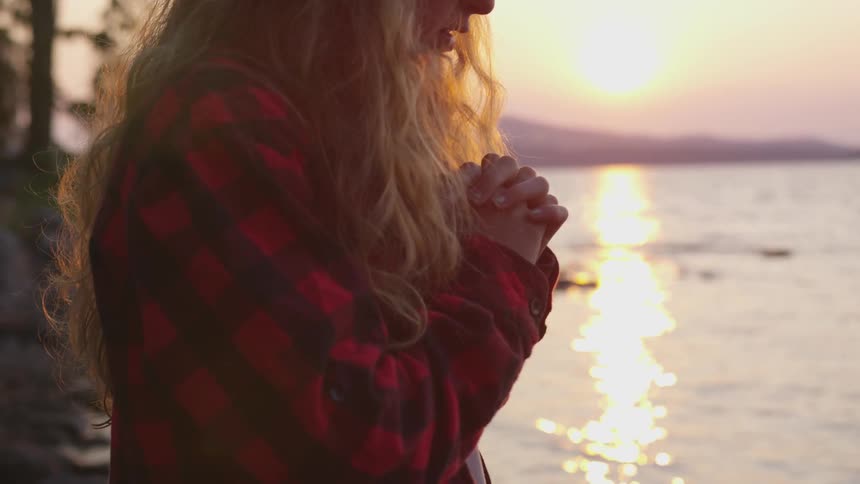 A blonde white woman praying while standing on the shore at sunset
