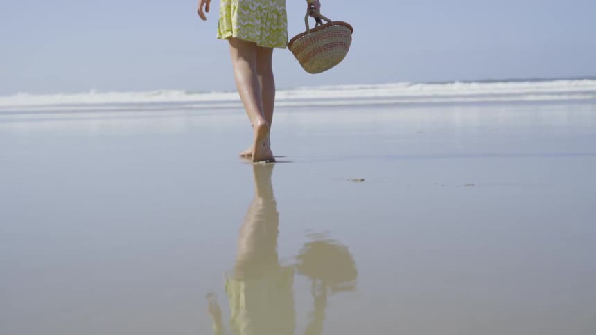 A barefoot white female in a yellow dress walking on the beach towards the water while carrying a wicker basket